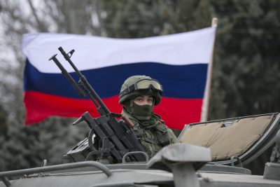 A pro-Russian man holds a Russian flag behind an armed servicemen on top of a Russian army vehicle outside a Ukrainian border guard post in the Crimean town of Balaclava